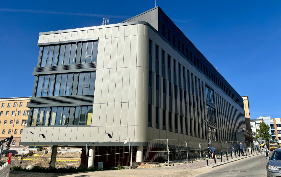 Ein modernes Bürogebäude mit hellgrauer und anthrazit-farbener Fassade ist von einem Bauzaun umgeben. Menschen bewegen sich auf dem Gehweg an dem Gebäude, auf der Straße fahren Autos und eine Radfahrerin.