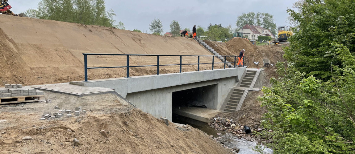 Eine Betonbrücke führt über einen Bach. Auf der Brücke ist ein Geländer. Auf der Treppe, die zur Straßen hinaufführt, gehen Bauarbeiter. Der Brückenhang besteht aus Sand. Ganz links steht ein Bagger auf der Brücke.