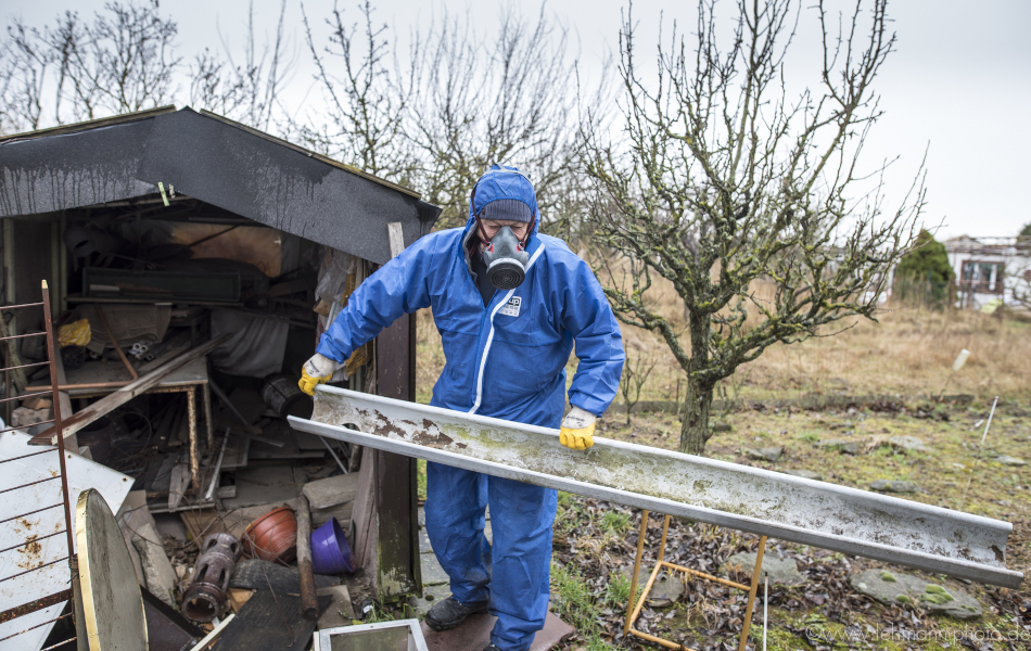 Ein Mann mit blauem Schutzanzug und Maske entfernt eine Regenrinne von einem maroden Gebäude, in dem sich zahlreiche alte Möbel und verrostete Gegenstände befinden.