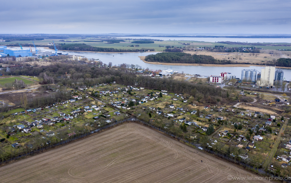 Blick von oben auf die Stadt Wolgast, die an einem Fluss, dem Peenestrom, liegt. Vor der Stadt liegt ein Feld, in der Stadt befinden sich mehre Kleingärten, Häuser, Bäume sowie Fabrik- und Werftgebäude am Flussufer.