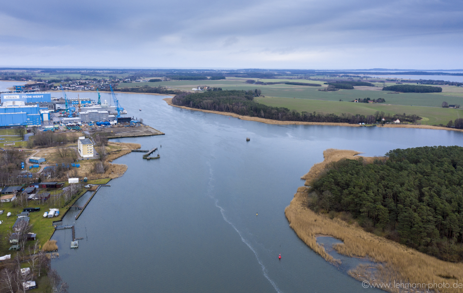 Blick über einen Fluss, dem Peenestrom: Links befinden sich Werftgebäude und rechts Felder. Im Hintergrund ist eine Ortschaft.