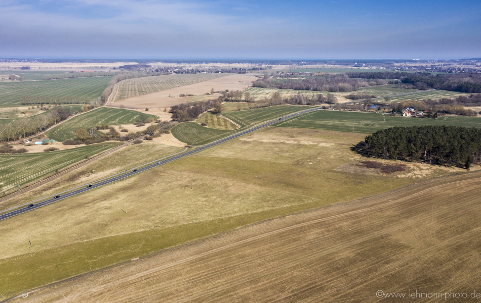 Blick über mehrere Felder, durch die eine Straße führt. Rechts liegen ein Wald und eine kleine Ortschaft. 