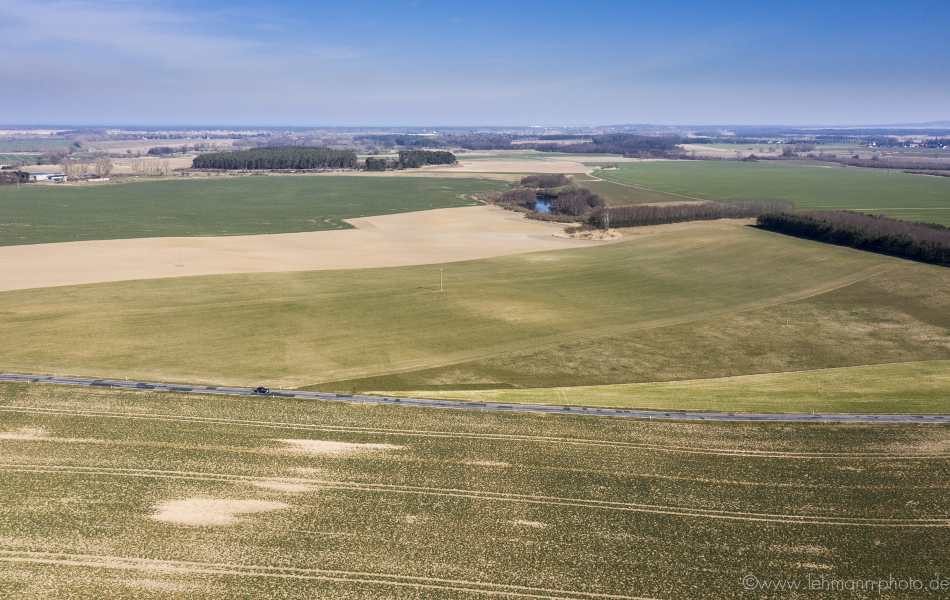 Blick über mehrere Felder und Wälder. Vorne führt eine Straße (die K 27) quer durch die Felder.