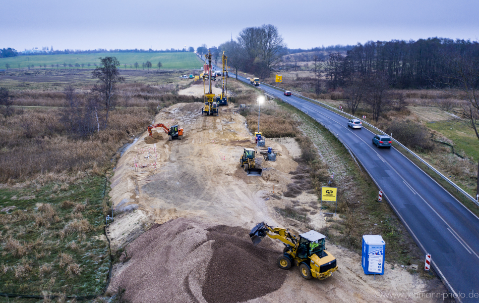 Blick auf eine Straße, auf der Lkw und Autos unterwegs sind. Es dämmert. Die Straße führt an Feldern und Bäumen vorbei und über einen Fluss, die Ziese. Links neben der Straße ist eine Baustelle am Fluss. Dort sind mehrere Bagger und Radlader aktiv.