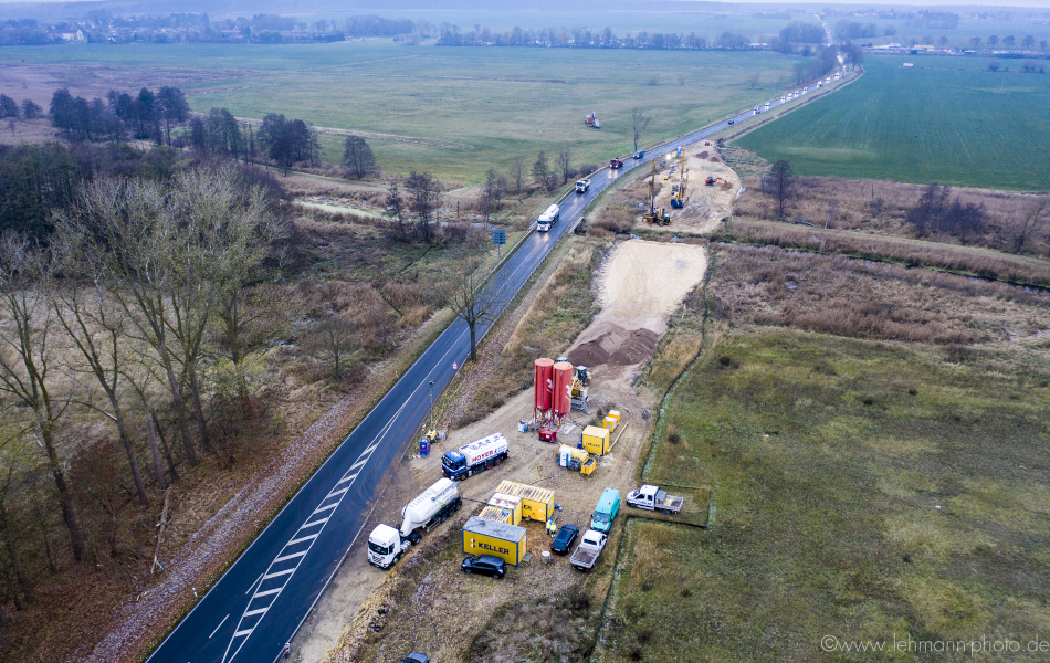 Blick von oben auf eine stark befahrene Straße mit Lkw und Autos. Die Straße führt an Feldern und Bäumen vorbei und über einen Fluss, die Ziese. Rechts neben der Straße ist eine Baustelle am Fluss. Dort stehen Lkw, Radlader und Bagger.  