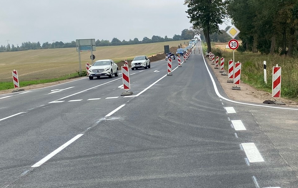 Auf einer Straße mit neuer Asphaltdecke und Fahrbahnmarkierung stehen mehrere Leitbaken in der Mitte und am Fahrbahnrand. Auf der linken Seite der Straße fahren Autos auf eine Kreuzung.