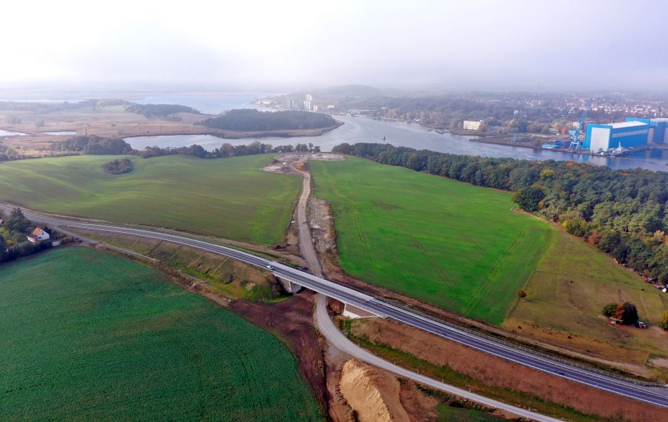 Eine Trasse aus Sand führt quer über ein Feld in Richtung eines Flusses. Die Trasse wird von einer neugebauten Straßenbrücke überführt und gekreuzt.