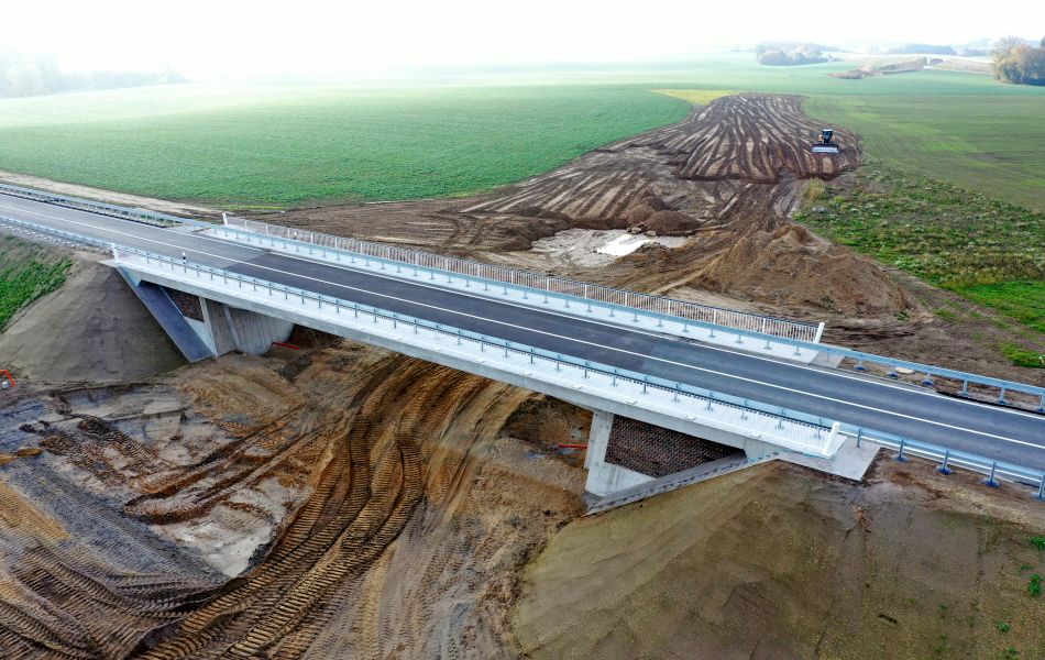 Eine Trasse aus Erde wird von einer neugebauten Straßenbrücke überführt und gekreuzt. Im Hintergrund bewegt ein Radlader Erde auf der Trasse.