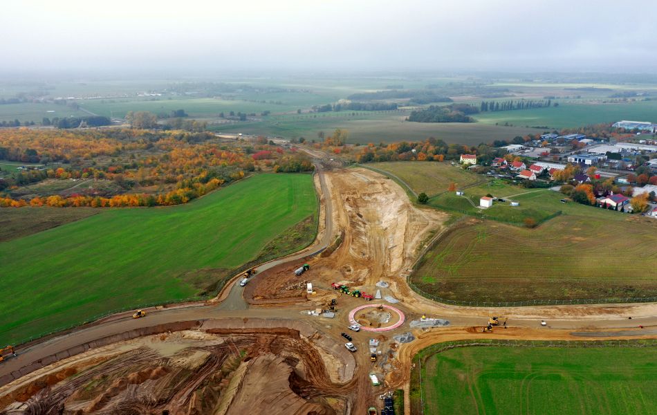  Auf einem Feld befindet sich eine riesige Baustelle, auf der Bagger, Radlader und Lkw Erde bewegen. Im Hintergrund ist ein Wäldchen und eine Stadt.