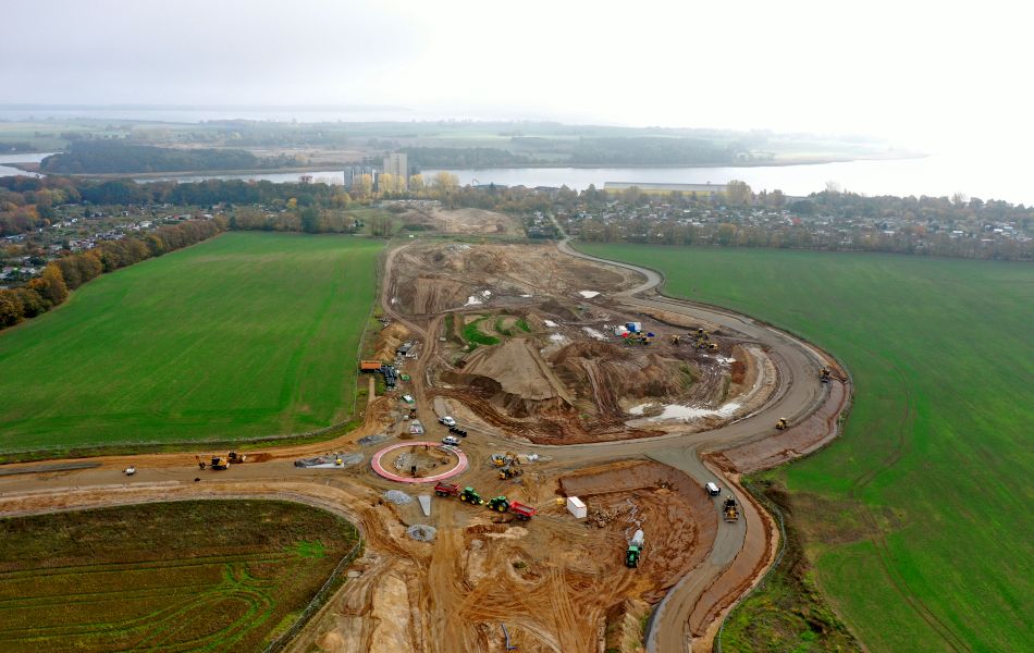 Auf einem Feld befindet sich eine riesige Baustelle, auf der Bagger, Radlader und Lkw Erde bewegen. Im Hintergrund sind eine Stadt, ein Fluss und Fabrikgebäude.