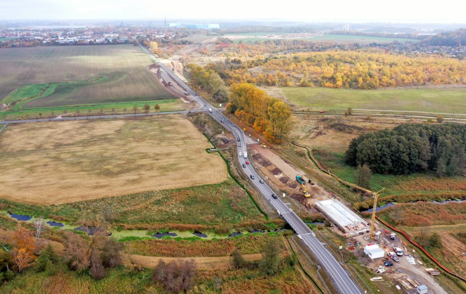 Auf einer Straße, die über einen Bach führt, fahren mehrere Autos. Direkt daneben befindet sich eine Baustelle mit einem hohen Kran, der an einem Fluss steht. Über den Fluss führt eine Brücke aus Beton. Im Hintergrund ist die Stadt Wolgast.