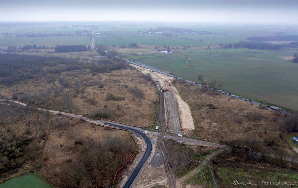 Eine Trasse aus Erde und teilweise Asphalt führt an Grünflächen und Bäumen vorbei und mündet in eine vielbefahrene Straße, der B 111. Im Hintergrund sind mehrere Felder.