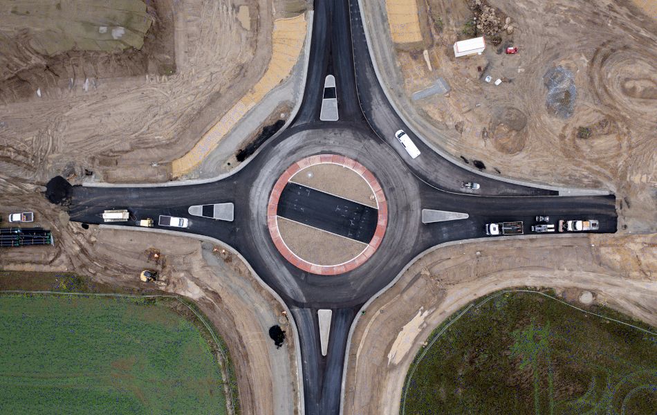 Blick von oben auf die Baustelle zur Errichtung des Kreisverkehrs an der Leeraner Straße. Baufahrzeuge sind auf dem frisch asphaltierten Kreisverkehr unterwegs. Neben dem Kreisverkehr befindet sich hauptsächlich Erde..