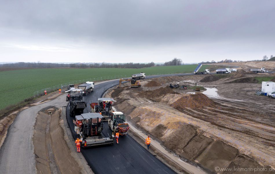 Mehrere Baufahrzeuge und Lkw verteilen Asphalt auf einer neu angelegten Straße, die sich auf einem Feld befindet.