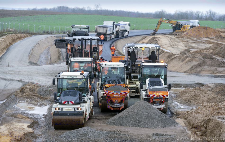 Mehrere Baufahrzeuge stehen auf einer Baustelle, auf der frischer Asphalt aufgebracht wird. Walzen, Radlader, Asphaltfertiger, Bagger und Lkw sind im Einsatz.