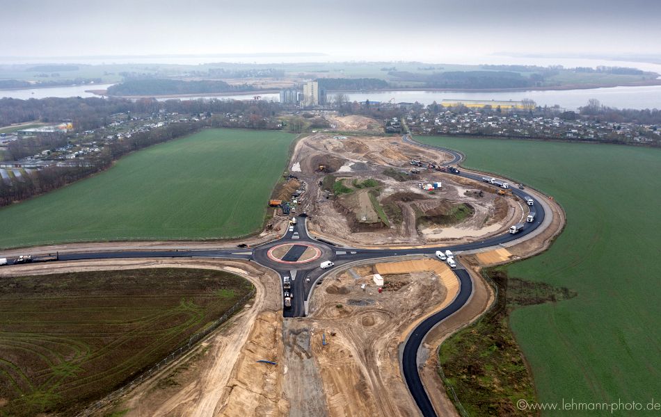 Auf einem Feld befindet sich eine riesige Straßenbaustelle, auf der zahlreiche Baufahrzeuge und Lkw Asphalt verteilen. Im Hintergrund sind eine Stadt, ein Fluss und Fabrikgebäude.