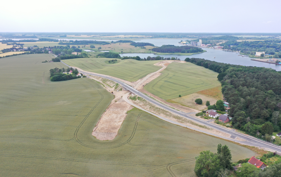 Eine Trasse aus Sand führt quer über ein Feld in Richtung eines Flusses. Die Trasse wird von einer neugebauten Straßenbrücke überführt und gekreuzt.