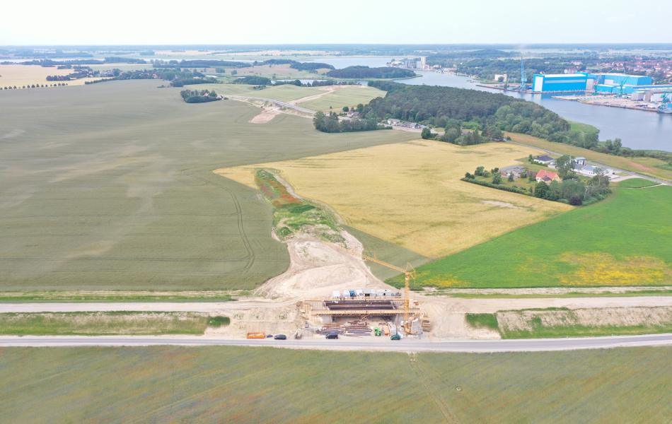 Auf einem Luftbild ist an einem Fluss ein blaues Werftgelände zusehen. Im vorderen Bereich steht ein riesiger Kran an einer Baustelle auf einem Feld. Von der Baustelle führt eine sandige Trasse einige Meter in Richtung einer Straßenbrücke mitten auf dem Feld. Auch hier ist eine sandige Trasse erkennbar.