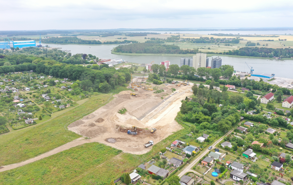 Zwischen zwei Wohnsiedlungen liegt eine große Baustelle, auf der Bagger, Radlader und Lkw Erde bewegen. Im Hintergrund sind ein Fluss und Fabrikgebäude.