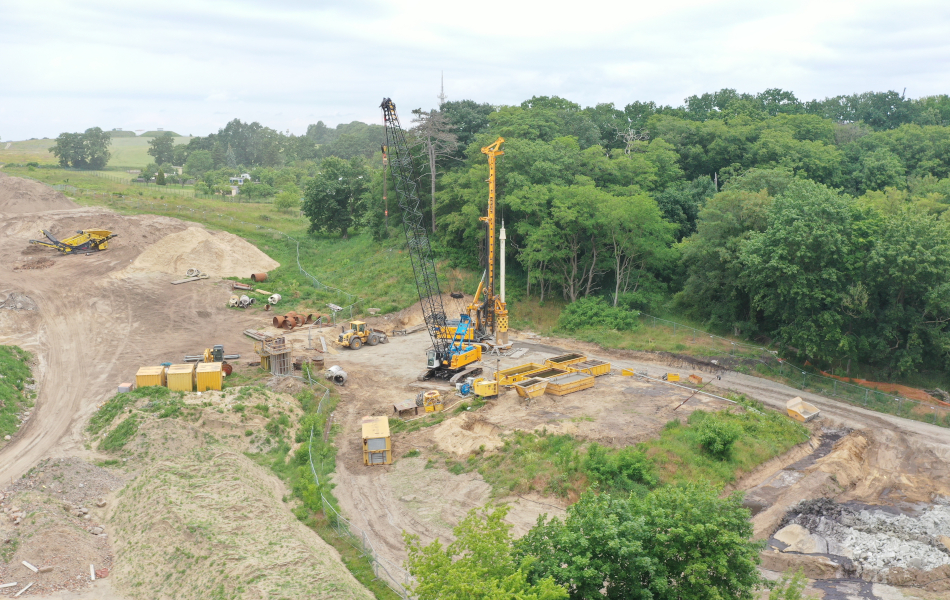 Ein hoher Kran und ein Bohrturm stehen auf einer Baustelle an einem Wäldchen.