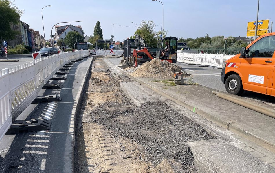 Auf einer mehrspurigen Straße stehen auf einer Baustelle ein Transport, ein Mini-Bagger und ein Lkw. In der Mitte der Straße ist eine langes Stück der Fahrbahn herausgefräst. Rechts davon befindet sich ein Sandhaufen.