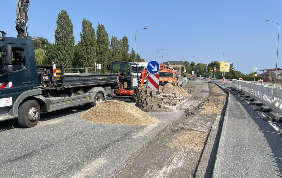 Auf einer mehrspurigen Straße stehen ein Lkw, ein Mini-Bagger und ein Transporter. In der Mitte der Straße ist eine langes Stück der Fahrbahn herausgefräst. Links davon befindet sich ein Sandhaufen.