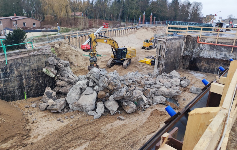 Auf einer Baustelle ist eine Wand durchbrochen werden. Schwere Betonreste liegen auf dem Boden. Im Hintergrund sind Bagger und eine Hubsteiger. 