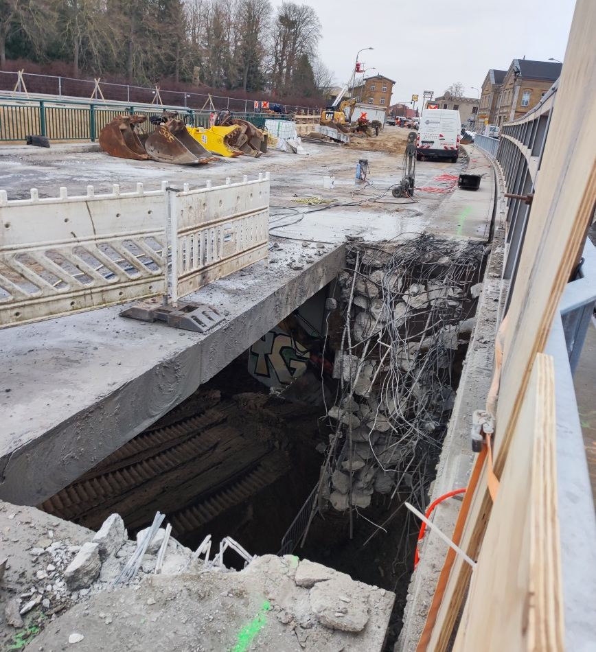 Auf einer Brückenbaustelle befindet sich ein offener Bereich, der mit Sicherheitsbaken abgesperrt ist, damit niemand in die Tief fällt. An einer Seite des offenen Bereiches hängen Reste des Überbaus in die Tiefe.