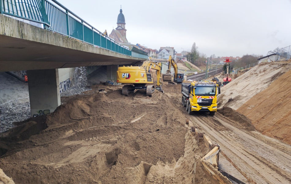 Zwei Bagger verteilen Kies auf einer Baustelle unter einer Brücke. Ein LKW transport den Kies zur Baustelle. Im Hintergrund sind Bahngleise, eine Stadt und ein Hubsteiger.