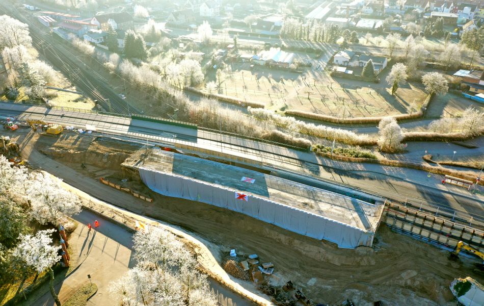 Blick von oben auf zwei parallel verlaufende Straßenbrücken, die über eine Bahnlinie führen. Die vordere Brücke ist von der bestehenden Straße abgekoppelt und von vorne mit einem Vorhang bedeckt.