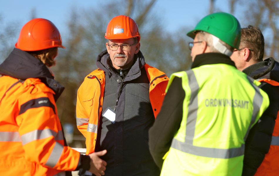 Drei Männer in orangener und gelber Schutzkleidung sprechen mit dem Minister, der ebenfalls eine Warnjacke und einen Helm trägt. 