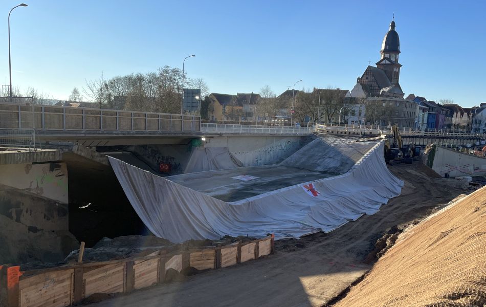 Eine Brücke ist eingestürzt. Ein Vorhang ist am Oberbau befestigt. Im Hintergrund ist eine weitere Brücke, die noch in Takt ist. Dahinter sind Häuser und eine Kirche. 