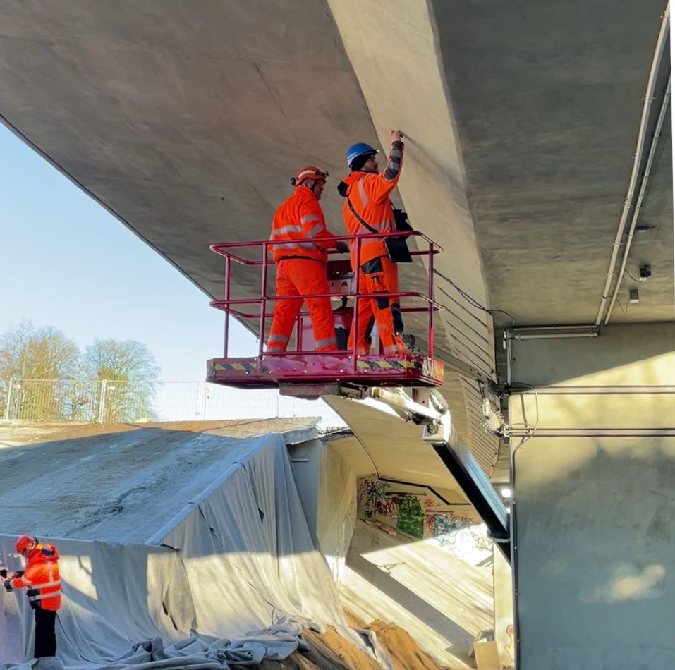 Zwei Ingenieure stehen in orangener Schutzkleidung und mit Helmen auf dem Kopf auf einem Hubsteiger unter einer Brücke. Sie begutachten die Unterseite der Brücke. Links daneben ist eine Brücke, die abgerissen und niedergeführt wurde.