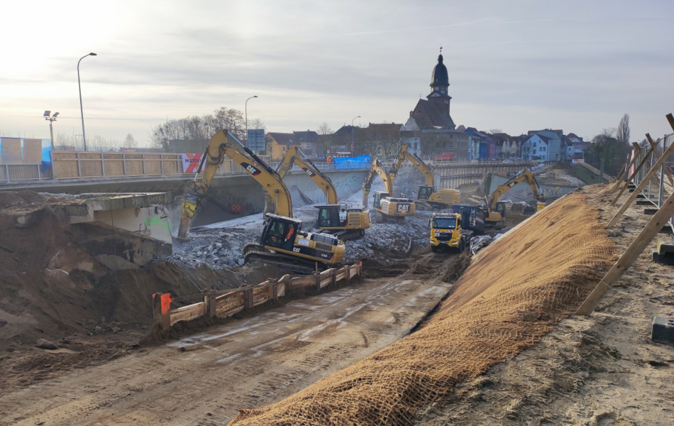 Mehrere Bagger tragen den Beton einer niedergeführten Brücke ab. Daneben befindet sich noch eine intakte Brücke, über die Autos fahren. Um die Bagger herum befindet sich reichlich Geröll und Kies. Im Hintergrund sind eine Kirche und Häuser.