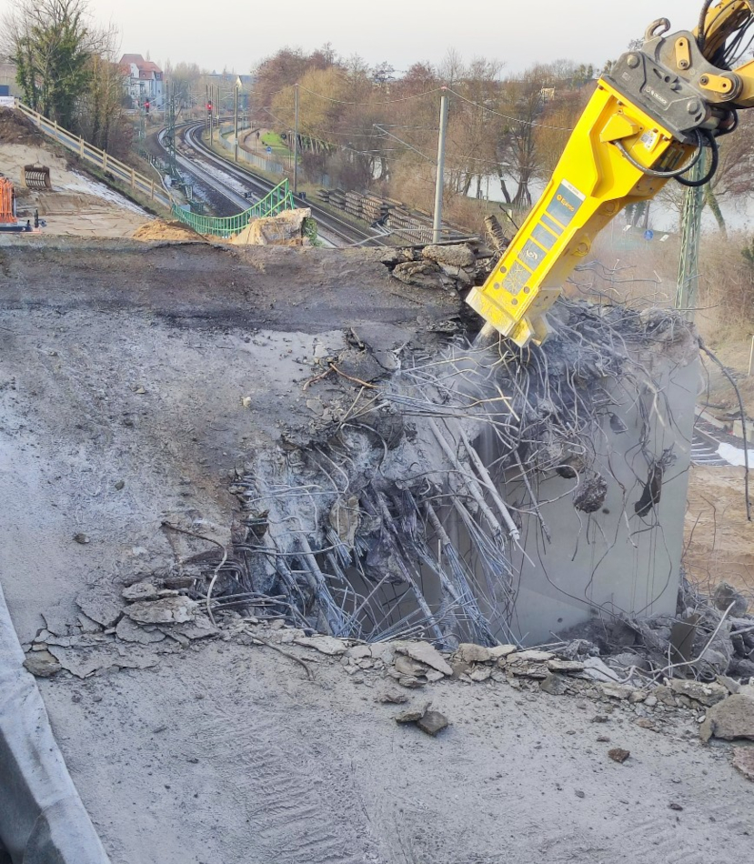 Mit schwerem Gerät, einem riesigen Bohrer, wird der Betonoberbau einer Brücke aufgebrochen und in kleine Stücke zerteilt. Im Hintergrund sind Bahnschienen und Häuser.