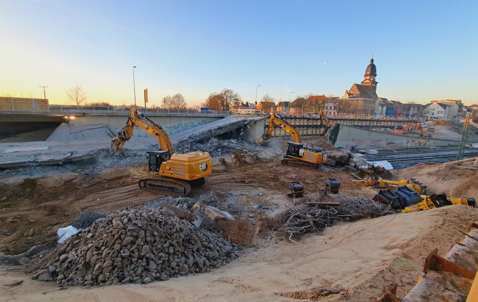 Zwei Bagger arbeiten auf einer Baustelle, während die Sonne aufgeht. Um die Bagger herum befindet sich reichlich Geröll und Kies. Im Hintergrund sind eine Kirche und Häuser.