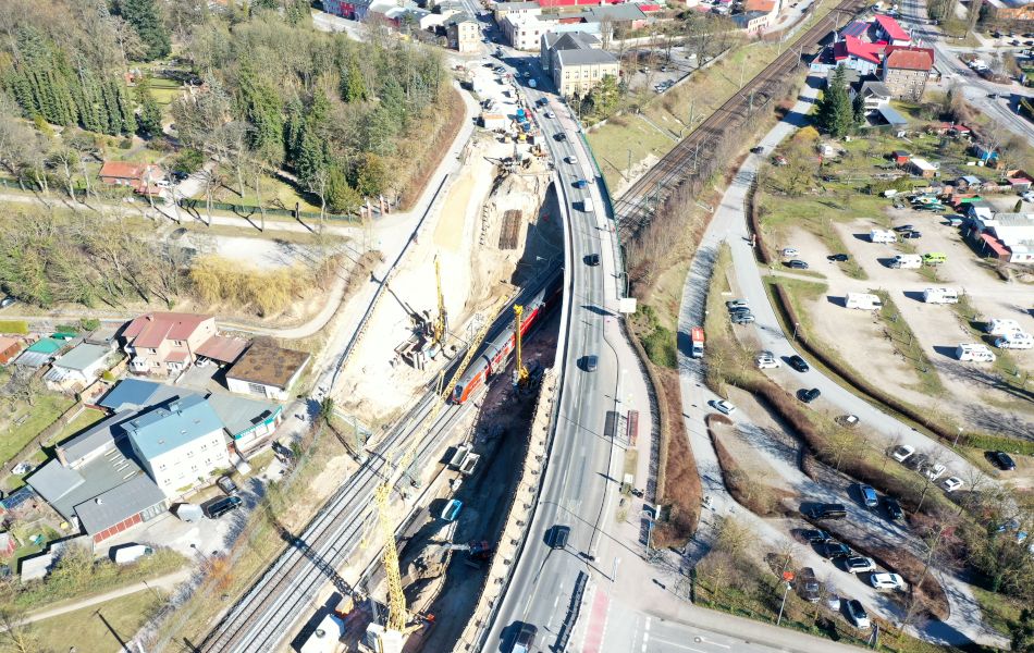 Auf einer Brücke fahren mehrere Autos durch eine Stadt. Die Brücke führt über Bahngleise, auf denen ein Zug fährt. An der Brücke stehen mehrere Baukräne. Im Umfeld der Brücke befinden sich Gebäude und Parkplätze.