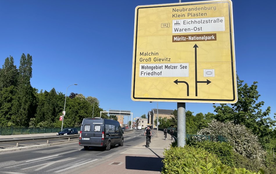 Eine vierspurige Straße führt über eine Brücke. In der Mitte der Straße befinden sich Leitplanken zwischen den Fahrbahnen. Radfahrer sind auf einem straßenbegleitenden Geh- und Radweg unterwegs. Ein großes gelbes Wegweiser-Schild am Straßenrand zeigt die Fahrrichtungen nach Neubrandenburg und Malchin.