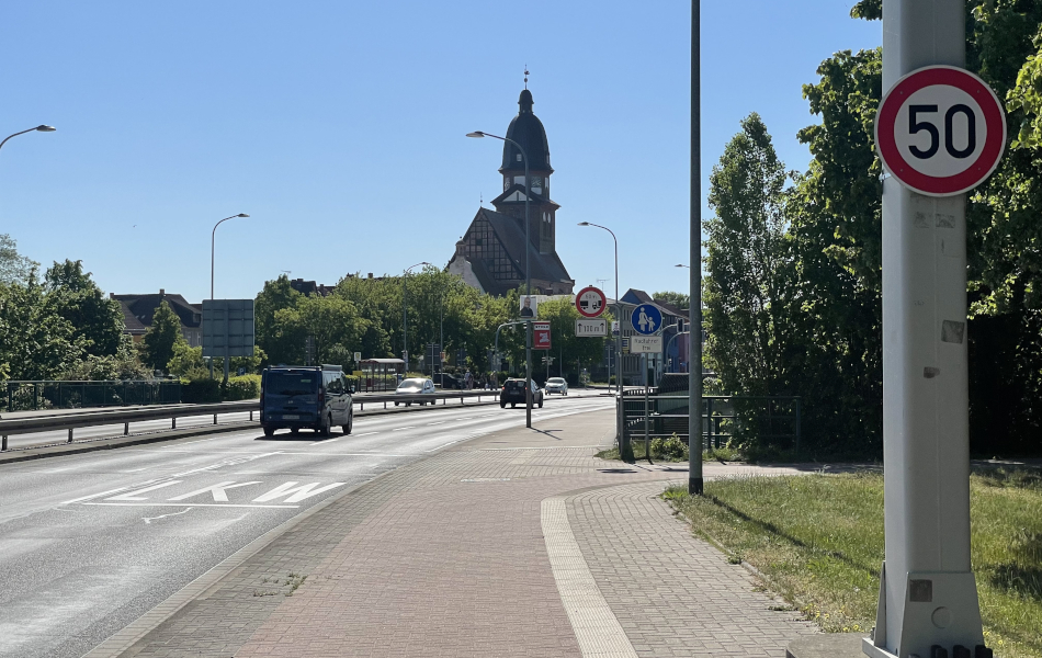 Eine vierspurige Straße mit Geh- und Radweg führt über eine Brücke. In der Mitte der Straße befinden sich Leitplanken zwischen den Fahrbahnen. Am Straßenrand steht ein Tempo-50-Schild.