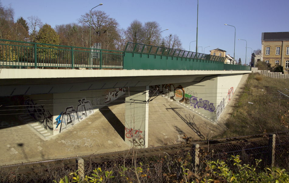 Bahngleise führen unter einer Straßenbrücke hindurch. Rechts von den Gleisen ist ein begrünter Hang und im Hintergrund sind Häuser