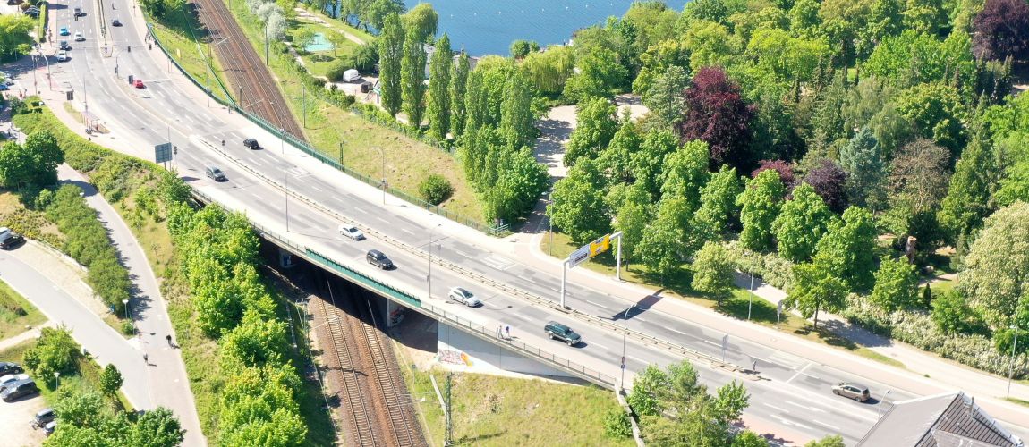 Autos fahren auf einer mehrspurigen Straßenbrücke, die über Bahngleise führt. An den Seiten der Straße befinden sich ein Parkplatz, Bäume und ein See.