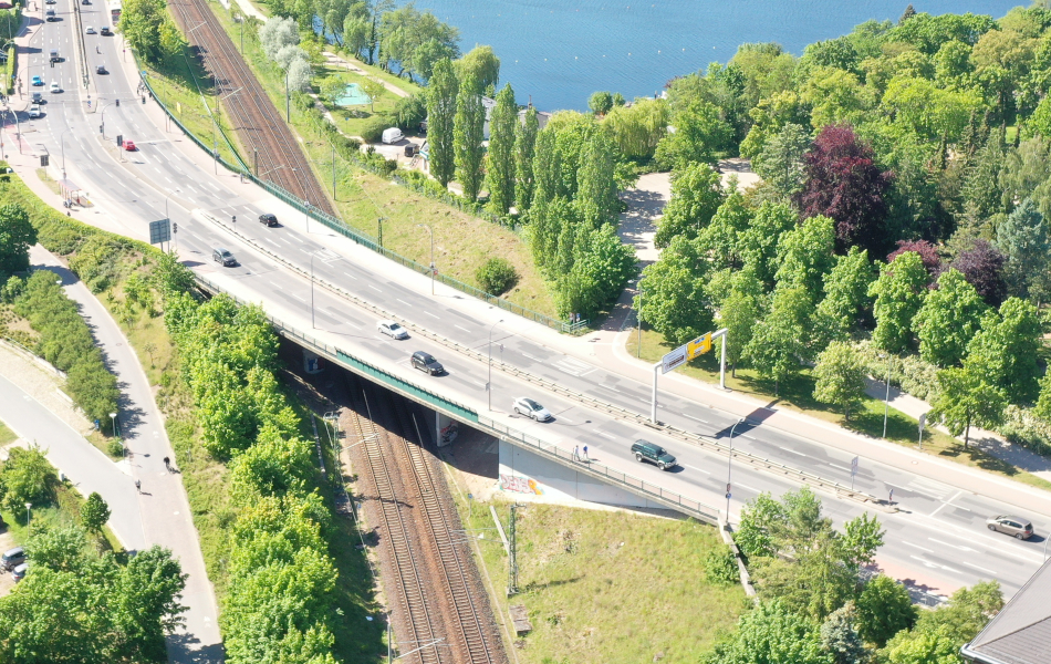 Autos fahren auf einer mehrspurigen Straßenbrücke, die über Bahngleise führt. An den Seiten der Straße befinden sich ein Parkplatz, Bäume und ein See.