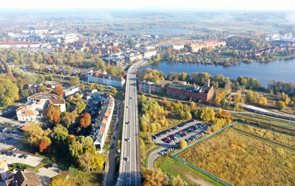 Mehre Autos fahren über eine Straßenbrücke, die durch ein Stadtgebiet und über ein Gewässer führt. Neben der Brücke sind Wohnhäuser und Parkplätze.