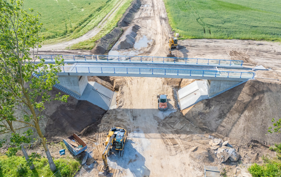 Eine in Bau befindliche Brücke mit blauem Geländer überführt einen Sandweg. Unter der Brücke fahren zwei Bagger und ein Radlader.