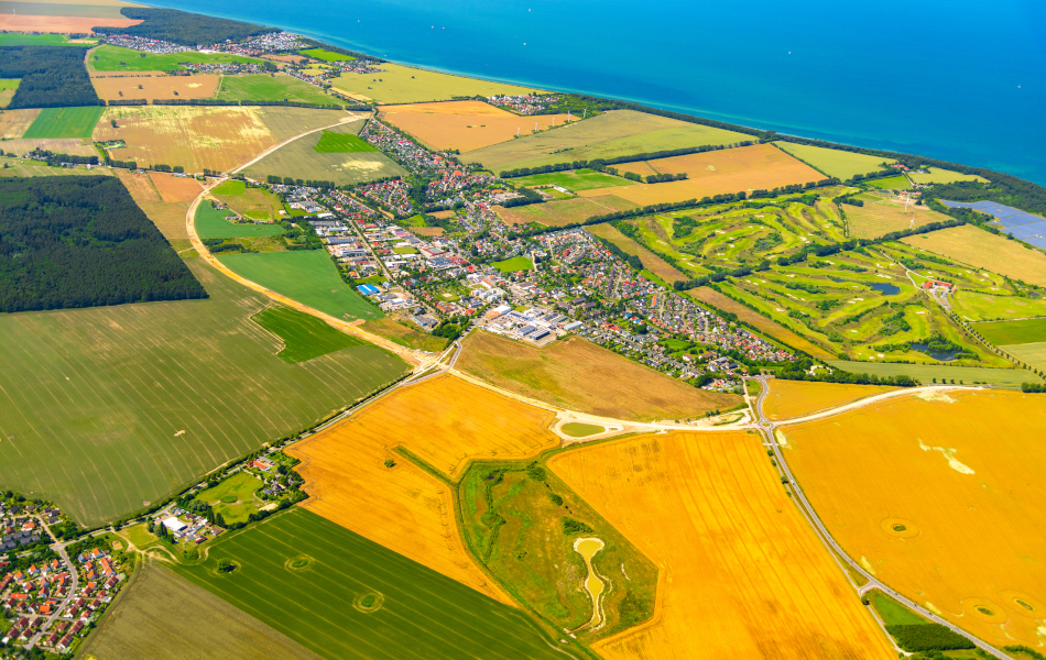 Auf einer Luftaufnahme ist der Ort Elmenhorst, eingebettet in einer Felderlandschaft an der Ostsee, abgebildet. In einem Halbkreis liegt die neue Ortsumgehungstrasse, überwiegend aus Sandboden bestehen, um den Ort herum.