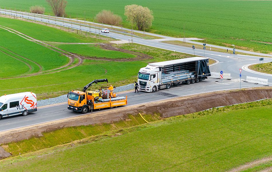 Ein Lkw liefert Material für den Aufbau von Schutzplanken an. Davor steht ein Baufahrzeug mit einem Kran, um das Material an der neuen Straße zu verteilen. 