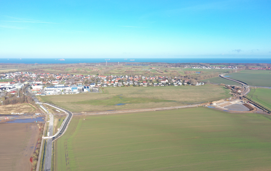 Luftaufnahme: Über die Felder vor der Ortschaft Elmenhorst führt eine neue Trasse, an der auf der rechten Seite ein Becken liegt. Links führt eine asphaltierte Straße, die einen kleinen Knick macht, über die neue Trasse nach Elmenhorst. Im Hintergrund ist die Ostsee.