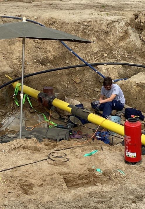 Leitungen, die unter der Erde entlang führen, sind freigelegt. Unter dem Schutz eines Sonnenschirmes arbeitet ein Techniker an einer Leitung.