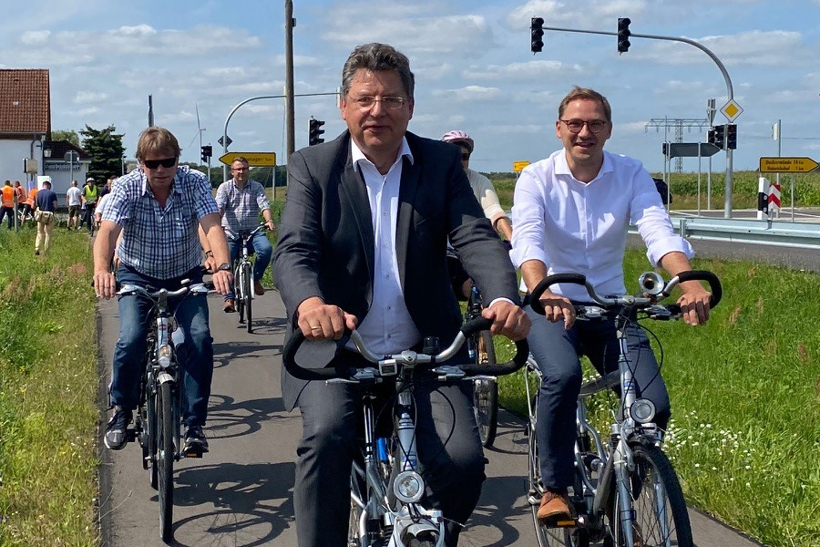 Drei Männer fahren Fahrrad auf einem neuen Radweg an einer Straße. Im Hintergrund sind weitere Radfahrer, eine Kreuzung mit Ampeln und ein Gebäude.