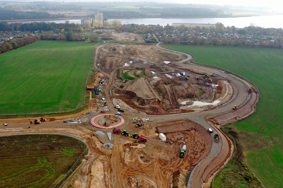 Auf einem Feld befindet sich eine riesige Baustelle, auf der Bagger, Radlader und Lkw Erde bewegen. Im Hintergrund sind die Stadt Wolgast, der Fluss Peene und Fabrikgebäude.